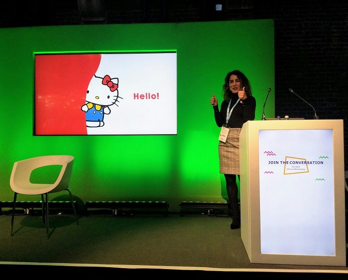 Martina Longueira gives a thumbs up on the Festival of Marketing content stage in front of a picture of Hello Kitty saying hello