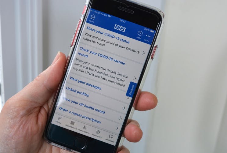 Stock photograph of a hand holding a smartphone with the NHS app open on it listing options to share your Covid-19 status, check your vaccine record, order a repeat prescription, and more.