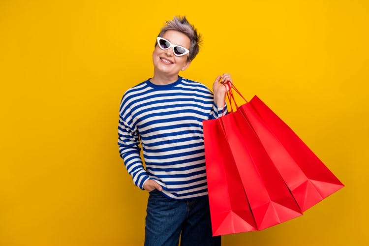 white woman with short blonde hair wearing white sunglasses and navy and white striped t-shirt smiles holding three red paper shopping bags against a yellow background